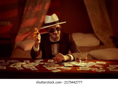 A joyful gambling man in elegant suit, hat and sunglasses sits in a bedroom at a table playing cards for money and smoking a cigar. Retro style. Mafia, gambling and the criminal world. - Shutterstock ID 2173442777