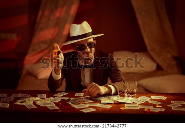 A\
joyful gambling man in elegant suit, hat and sunglasses sits in a\
bedroom at a table playing cards for money and smoking a cigar.\
Retro style. Mafia, gambling and the criminal\
world.