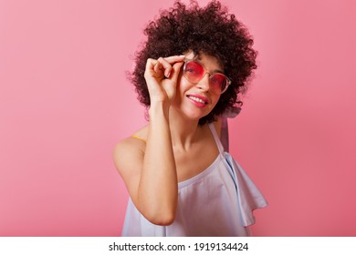 Joyful girl in blue summer blouse holding her pink glasses on pink background. Attractive woman smiling and posing on isolated backdrop. High quality photo