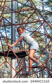 Joyful girl climbs a rope structure at a park, her smile expressing the thrill of adventure amidst tall pines, perfect for themes of active childhood, and sports activities. High quality photo