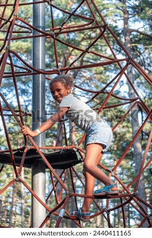 Joyful girl climbs a rope structure at a park, her smile expressing the thrill of adventure amidst tall pines, perfect for themes of active childhood, and sports activities. High quality photo