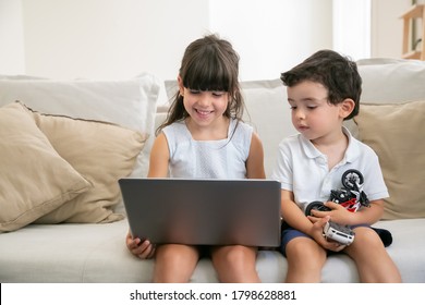 Joyful Girl And Her Little Brother Sitting On Couch At Home, Using Laptop For Video Call, Online Chat, Watching Video Or Movie. Front View. Kids Leisure Time Or Internet Concept