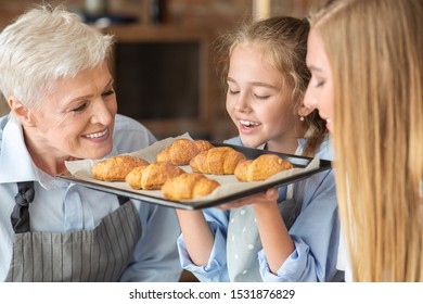 Joyful Girl Holding Tray And Smelling Freshly Baked Croissants, Cooking With Mom And Granny, Kitchen Interior, Close Up