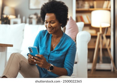 Joyful glad African American woman in a casual sits on the floor engaged with her smartphone at home, enjoy chatting. African woman using phone sitting on floor - Powered by Shutterstock