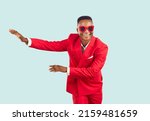 Joyful good looking African man in trendy outfit dancing in modern fashion studio. Happy handsome young Tanzanian guy wearing funky red suit and cool sunglasses dancing isolated on blue background
