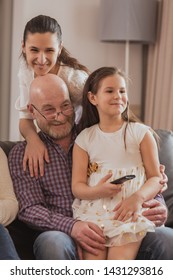 Joyful Grandfather In Glasses Relaxing With Grandchildren At Home. Happy Man Sitting With Lovely Girl On Sofa And Watching Tv While Daughter Standing Behind And Looking At Camera. Concept Of Family.