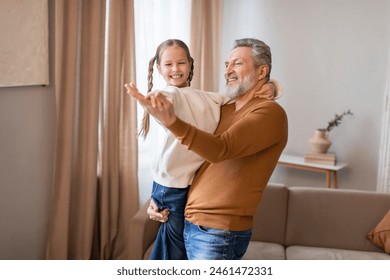 A joyful grandfather is holding his granddaughter as they dance in a warm and inviting living room. They are both smiling, sharing a moment of happiness and bonding while the sunlight filters in - Powered by Shutterstock