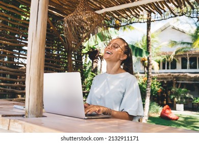 Joyful graphic designer in eyeglasses using netbook computer during remote working outdoors, cheerful female copywriter with laptop laughing and rejoicing - digital nomad and freelance lifestyle