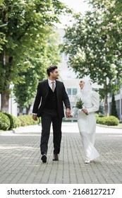joyful groom holding hands with muslim bride with wedding bouquet and walking together