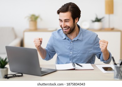 Joyful Guy At Laptop Computer Shaking Fists Gesturing Yes Celebrating Business Success Sitting In Modern Office. Great News And Victory Celebration Concept