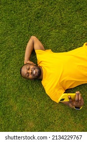 Joyful handsome african american male laying on the grass, relaxing and taking a break after workout. Smiling happy man using smartphone, talking with friends by video call outdoors.