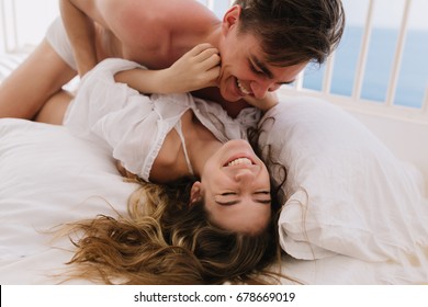 Joyful handsome boy in underwear tickles his laughing long-haired girl, lying with eyes closed. Portrait of cute young couple embracing on white bed and having fun on weekend together