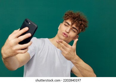 A Joyful, Handsome Man With Curly Hair Stands Against A Green Background And Holds His Black Smartphone In His Hand, Blowing A Kiss