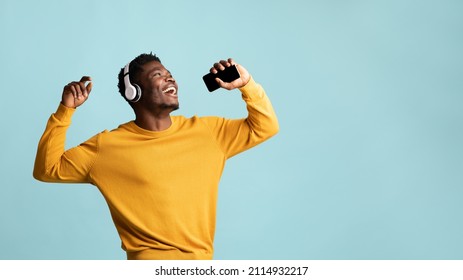 Joyful handsome millennial black guy in yellow cardigan singing karaoke, using wireless stereo headphones and modern smartphone as microphone, blue studio background, panorama, copy space