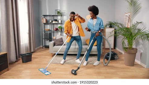 Joyful Happy African American Young Married Couple Wife And Husband Cleaning Living Room And Dancing, Guy Mopping And Woman Vacuuming Floor Having Fun Making Funny Rhythmic Moves In Cozy Modern Home