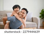 Joyful and happy Asian dad playing with his little son in the living room, piggyback, spending fun time together at home.