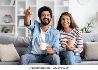 Joyful happy beautiful young indian lovers sitting on couch in living room, watching movie or tv, drinking coffee at home, cheerful hindu man pointing at camera, copy space