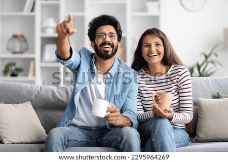 Joyful happy beautiful young indian lovers sitting on couch in living room, watching movie or tv, drinking coffee at home, cheerful hindu man pointing at camera, copy space