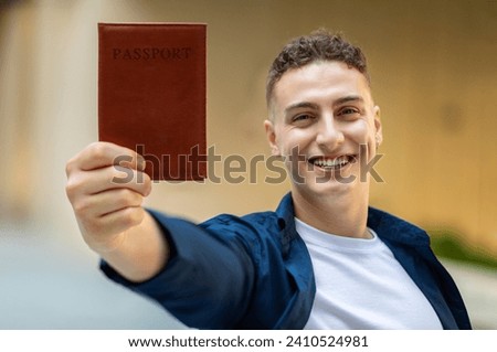 A joyful happy european young man holding up a passport with a brown cover towards the camera, with a big smile, wearing a navy shirt and a white t-shirt in city, close up