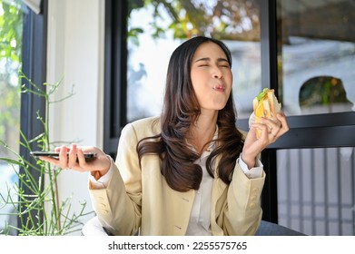 Joyful, happy and hungry millennial Asian businesswoman enjoying with her healthy breakfast, eating a sandwich at the cafe. happiness overload from yummy food - Powered by Shutterstock