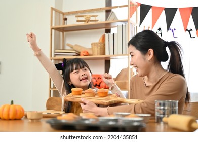 Joyful And Happy Little Asian Girl In Halloween Costume Having A Great Time With Her Mom, Baking Halloween Cupcakes For The Halloween Party With Her Mom.