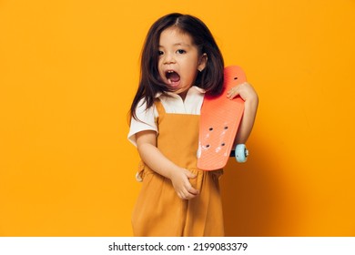 Joyful, Happy, Little Girl Of Preschool Age In An Orange Sundress Stands On An Orange Background With Her Mouth Wide Open With Delight Holding A Small Skateboard In Her Hand.