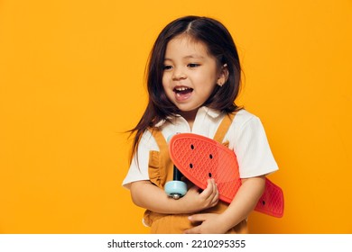 Joyful, Happy, Little Girl Of Preschool Age In An Orange Sundress Stands On An Orange Background With Her Mouth Wide Open With Delight Holding A Small Skateboard In Her Hand.