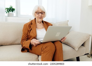 A Joyful, Happy Old Lady In A Brown Suit Is Sitting On A Beige Sofa Having Fun Communicating With Loved Ones Via Video Link Holding A Laptop On Her Lap