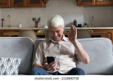 Joyful Happy Older 70s Cellphone User Man Waving Hand Hello At Gadget Frontal Camera, Speaking On Video Call, Online Conference Chat, Talking To Family, Laughing, Resting On Home Couch