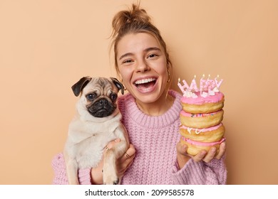Joyful happy woman enjoys birthday party celebration poses with pug dog and delicious festive donuts wears knitted sweater laughs out gladfully isolated over beige background. Pet anniversary