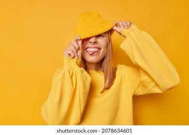 Joyful happy woman hides eyes with hat sticks out tongue has playful expression teases someone wears casual jumper isolated over yellow background squints happily shows true emotions. People and fun - Shutterstock ID 2078634601