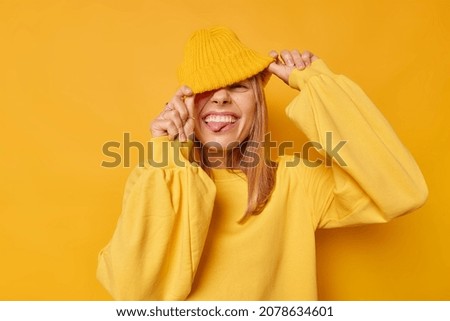 Joyful happy woman hides eyes with hat sticks out tongue has playful expression teases someone wears casual jumper isolated over yellow background squints happily shows true emotions. People and fun