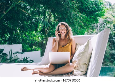 Joyful and happy woman sitting with laptop on white sofa next to pillow and speaking on phone while enjoying rest near pool and looking at camera - Powered by Shutterstock