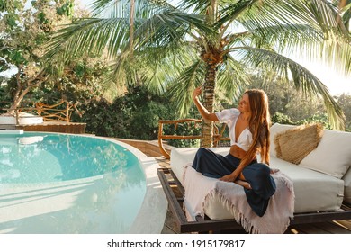 Joyful and happy woman sitting on white sofa under palm tree near swimming pool and making selfie photo on mobile phone