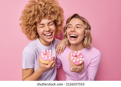 Joyful happy women take delicious doughnuts with marshmallow laugh gladfully do not care about calories enjoy eating dessert foolish around isolated over pink background. Unhealthy food concept