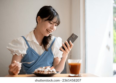 A joyful and happy young Asian woman is enjoying eating a croissant waffle and chatting with her friends online while relaxing in a cafe. Lifestyle concept - Powered by Shutterstock