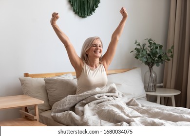 Joyful healthy older woman sitting on bed, waking up after good night rest on comfortable orthopedic mattress, rising up arms and stretching back. Happy mature grandma feeling energetic in morning.