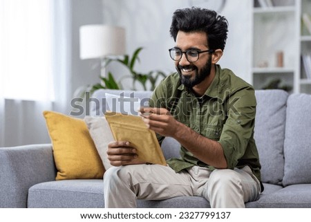 Joyful hispanic received letter mail notification man sitting at home in living room on sofa holding envelope smiling reading.