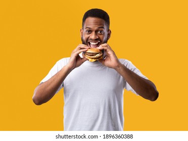 Joyful Hungry African Guy Eating Burger Smiling To Camera Posing Standing On Yellow Background. Studio Shot. Junk Food Eater Enjoying Cheeseburger. Fastfood And Cheat Meal