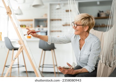 Joyful imaginative artistic elderly woman painting easel in leisure time sitting rope swing in living room at home 