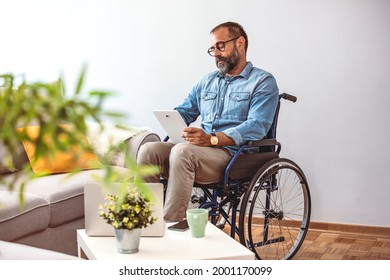 Joyful Impaired Mature Man In Wheelchair Using Tablet Computer At Home. Positive Retired Male With Physical Disability Browsing Web On Touch Pad, Watching Video Online Indoors