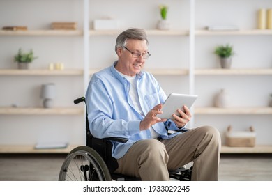 Joyful Impaired Senior Man In Wheelchair Using Tablet Computer At Home. Positive Retired Male With Physical Disability Browsing Web On Touch Pad, Watching Video Online Indoors