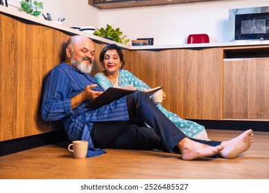 Joyful Indian Asian senior couple sitting comfortably on the kitchen floor, relaxing with cups of tea,fondly looking through an old photo album, reliving cherished memories together - Powered by Shutterstock