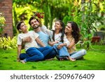 Joyful Indian couple with their kids sitting in a park, embracing each other and admiring a toy plane held by their daughter, symbolizing love, family time, imagination, and togetherness, aspiration