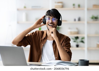Joyful indian guy in casual with wireless headset sitting at workdesk in front of modern laptop, singing karaoke, relaxing while working at office, copy space. Stress relief at job concept