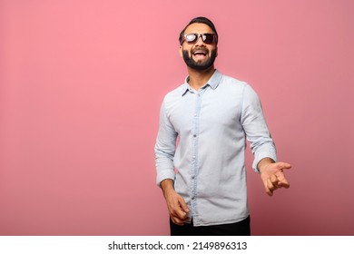 Joyful Indian man in sunglasses pretends guitar player, excited hispanic guy in casual jeans shirt enjoys playing on imaginary guitar, has a fun, isolated on pink