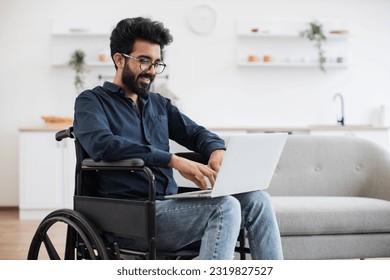 Joyful indian man in wheelchair holding laptop on knees while staying in middle of bright open-plan kitchen. Happy young adult surfing internet webpages while recovering after accident at home.