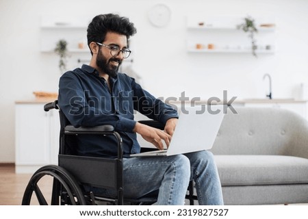 Joyful indian man in wheelchair holding laptop on knees while staying in middle of bright open-plan kitchen. Happy young adult surfing internet webpages while recovering after accident at home.