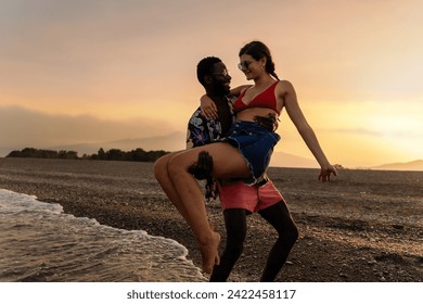 Joyful interracial couple embracing by the sea, sharing a light-hearted moment against a stunning sunset backdrop. - Powered by Shutterstock