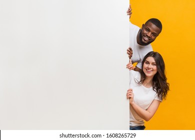 Joyful interracial couple peeking from behind white advertisement board with free space for your text, yellow studio background